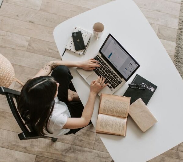 Mulher em uma mesa de café com laptop, xícara de café, smartphone com Instagram aberto e bloco de notas.